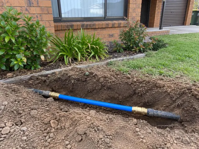 Open trench beside a brick house showing a repaired blue water pipe section in Marayong