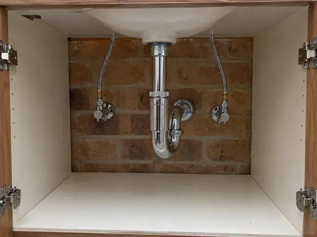 Open vanity cupboard with neat plumbing access panel and brick wall behind in Kings Langley